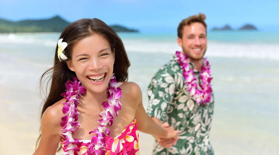 Happy couple holding hands on a Hawaiian beach, both wearing flower leis and smiling joyfully.