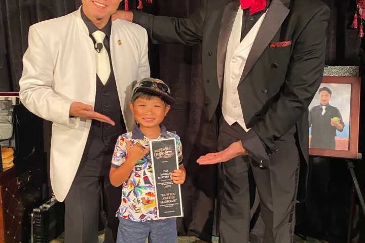 A child receiving a "Now You’re a Magician" certificate from two magicians in costume.