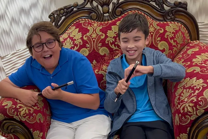 Two boys smiling and holding magic wands, sitting on an ornate red chair.