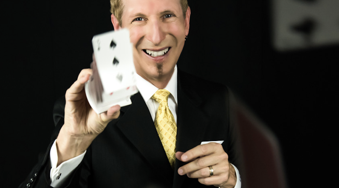 Magician performing a card trick, holding a deck of cards and smiling at the audience.