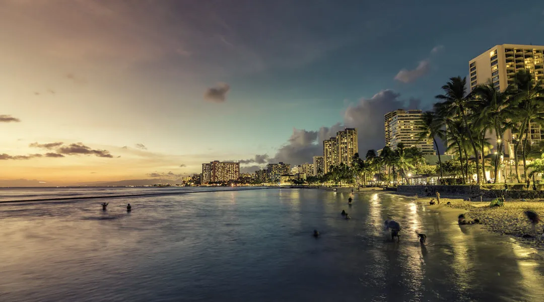 Waikiki Beach at dusk with city buildings and palm trees lining the shore, creating a serene tropical view.