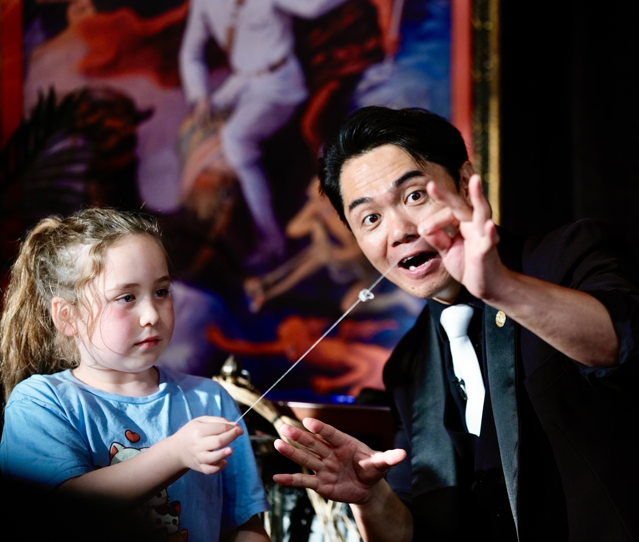 Magician performing a trick with a young girl, showing her how to make a magical gesture with a string.
