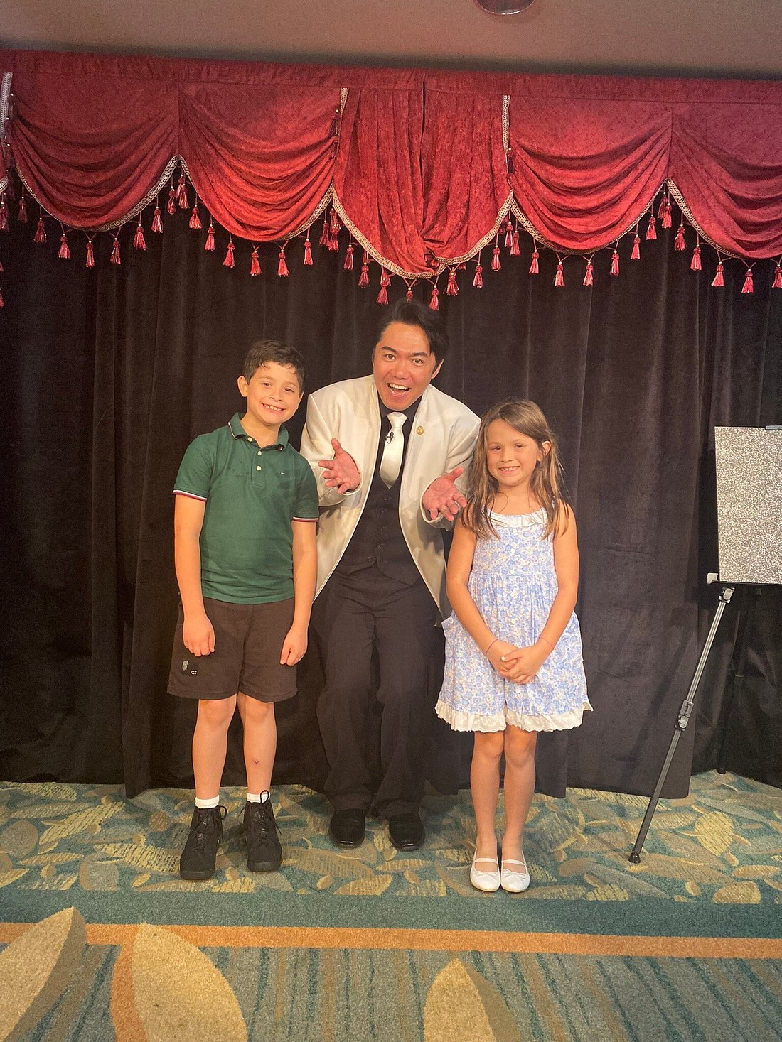 Magician performing with two children, one boy and one girl, on stage with red velvet curtains in the background during a live show.