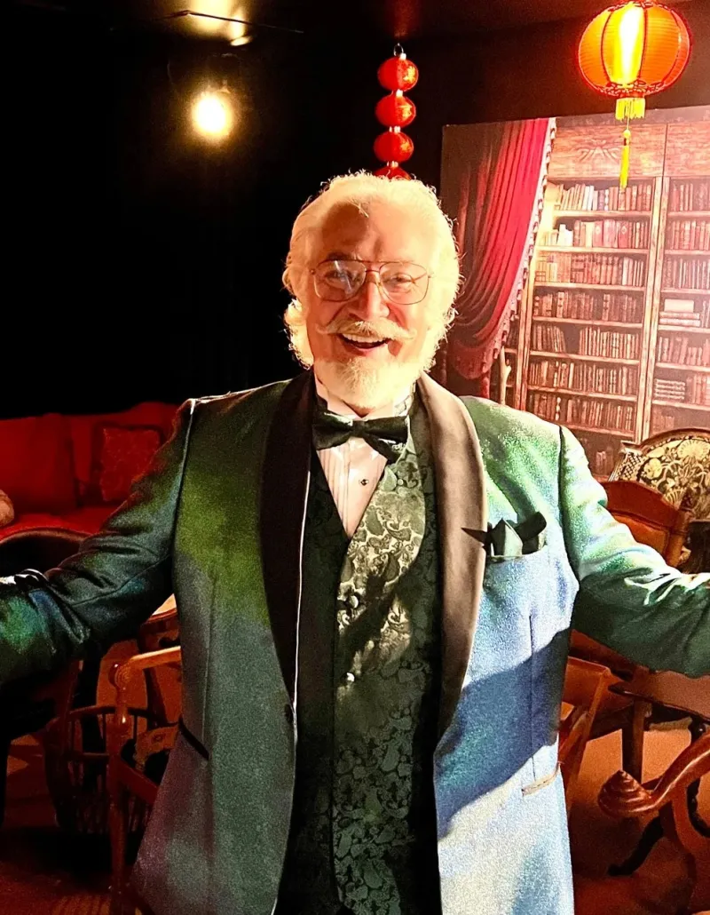Magician in a colorful tuxedo, smiling and welcoming the audience, with a cozy, magical backdrop of red lanterns and a vintage bookshelf.
