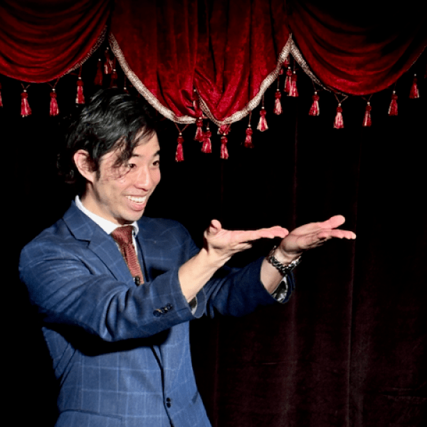Person in a suit gesturing with open hands under a red velvet curtain with tassels.