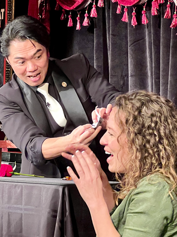 Magician performing a trick with a woman smiling on stage, vintage poster in background.