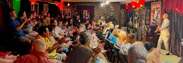 Audience clapping in a small, dimly lit theater with a performer on stage.